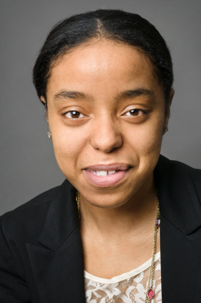 Headshot of a woman in her 20s who has heritage from Morocco with medium-light skin tone and black hair, parted and pulled back. She is wearing a black blazer, white lace top and a gold and jeweled necklace. She is smiling at the camera.