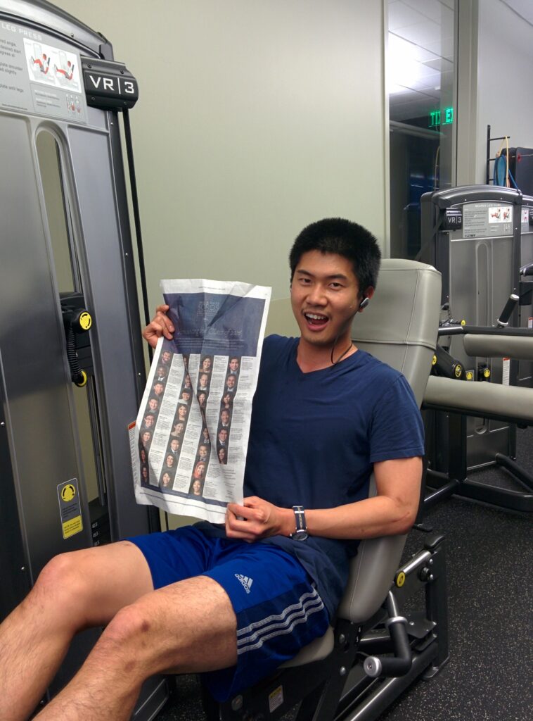 Photograph of a man in his 20s with medium skin tone and short black hair. He is wearing a blue t-shirt, blue shirts and sitting in a grey chair, holding up the New York Times.