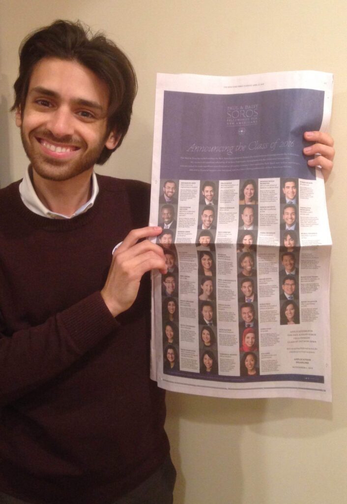 Photograph of a man in his 20s who has light skin tone and shaggy dark brown hair and a trim beard. He is wearing a maroon sweater, white button up underneath and holding the New York Times. 