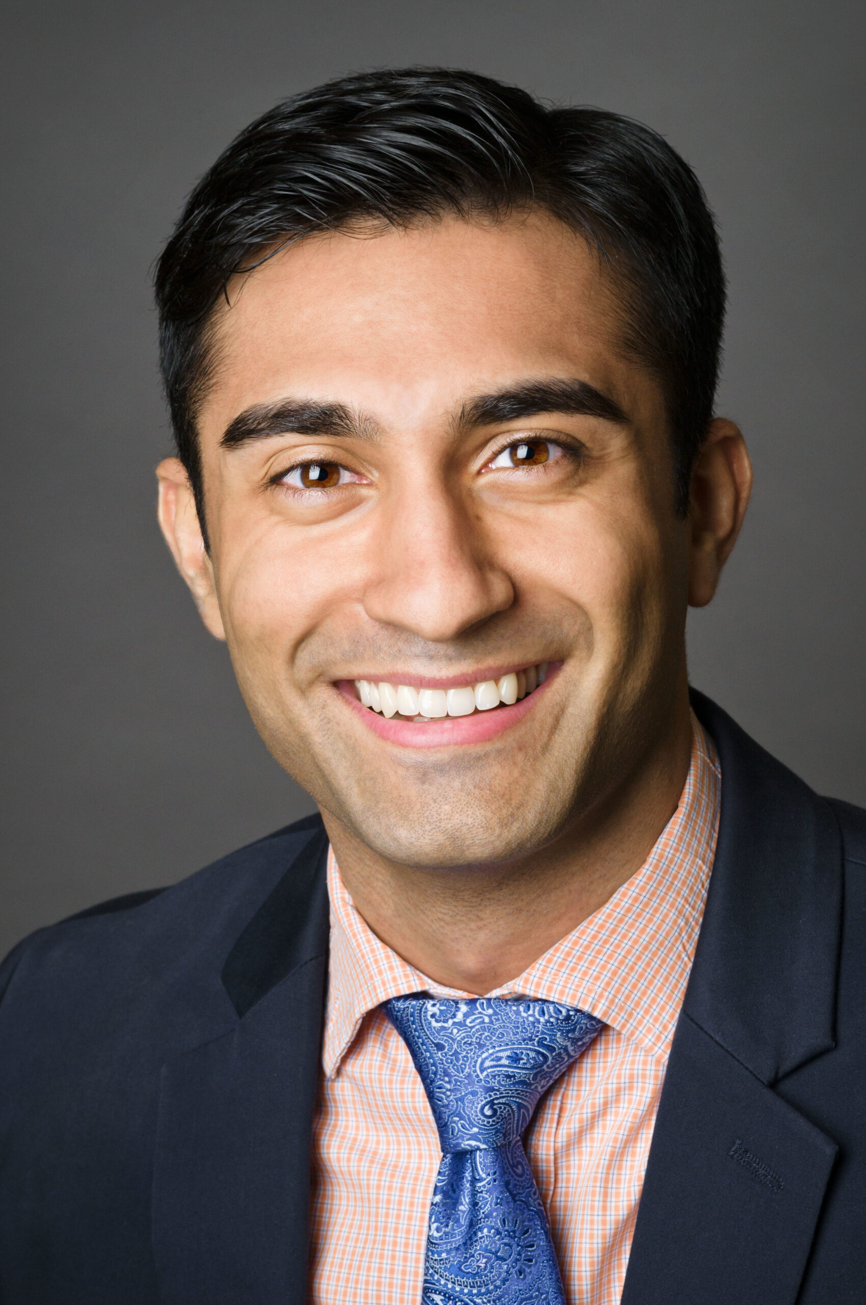 Headshot of a man in his 20s who has heritage from United Kingdom with light-medium skin tone and crew cut black hair. He is wearing a navy suit, orange and white gingham button up and blue paisley tie. He is smiling at the camera.