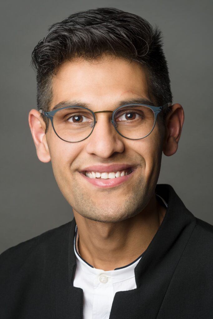 Headshot of a man in his 20s who has heritage from India, he has medium skin tone and crew cut black hair. He is wearing a black eastern collar suit and white shirt, he has thin framed round glasses. He is smiling at the camera. 