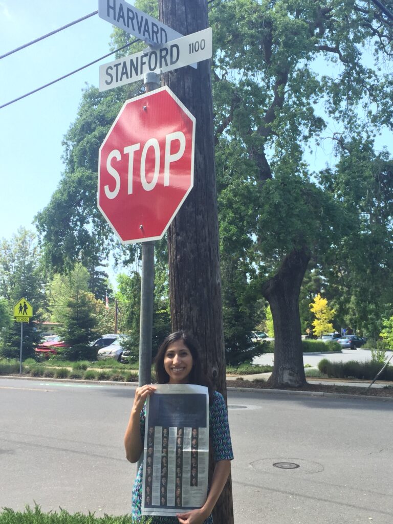 Photograph of a woman in her 20s with light skin tone and long black hair. She is standing next to a stop sign at the corner of "Harvard" and "Stanford" streets, and holding up the New York Times. 