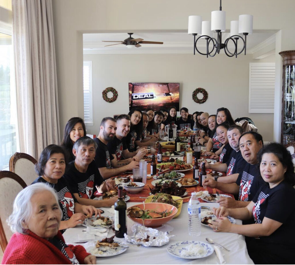 Photograph of a large group of people seated around a table. The table is full of food, the table is so long the people at the far end can barely be seen. 