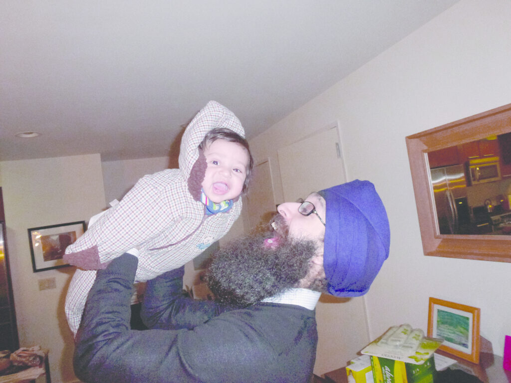 A man in his 30s, wearing a jacket and purple turban with an expression of joy on his face, lifts a child wearing a tartan onesie into the air, the child is also laughing.