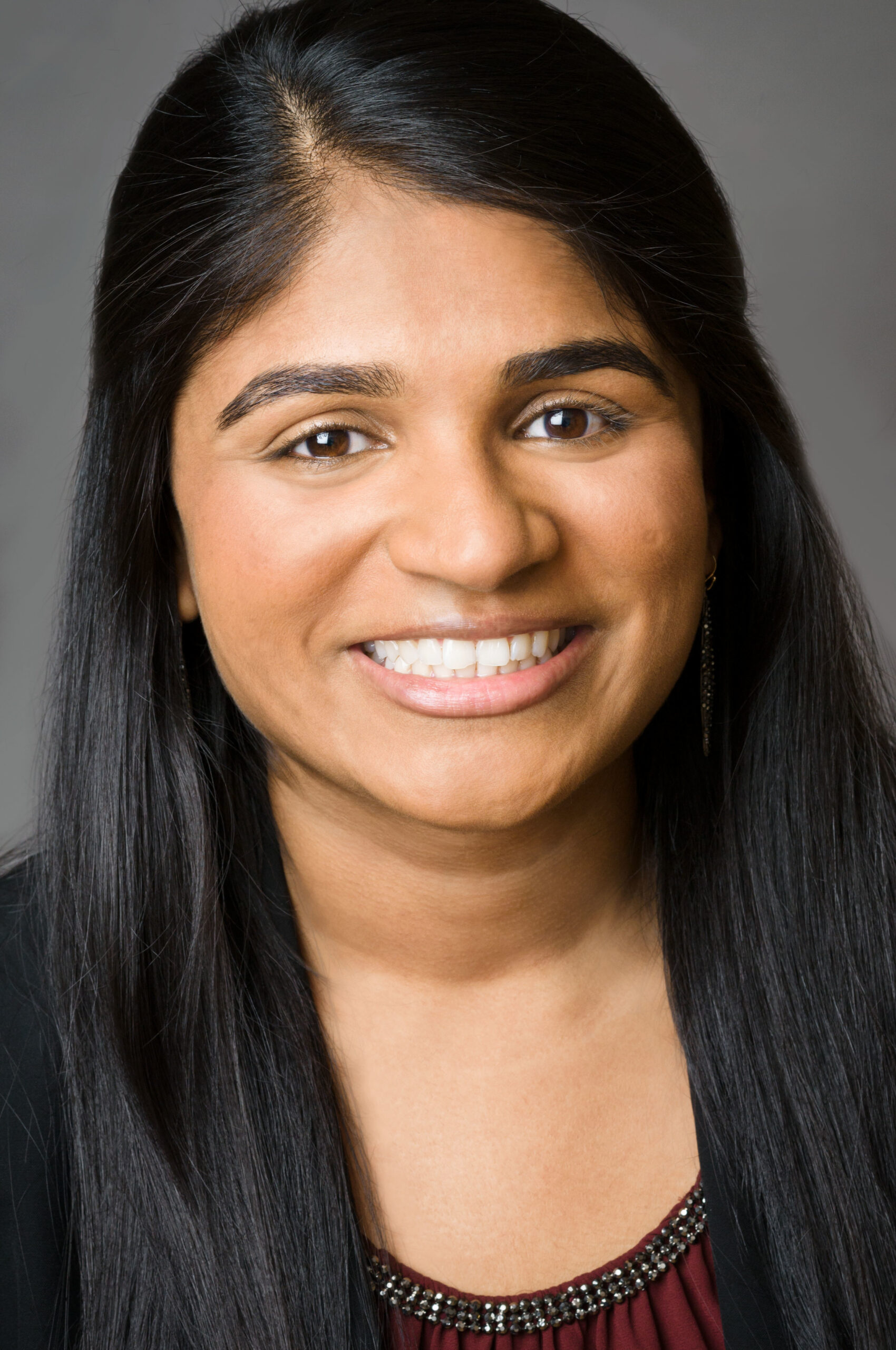 Headshot of a woman who has heritage from India with medium skin tone and long black side parted hair pulled half back. She is wearing a black blazer, and red blouse with bejeweled neck line. She is smiling at the camera. 