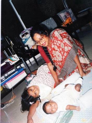 Old photograph of three people. One woman, the oldest person in the picture, is wearing a red saari and sitting. Next to her a young girl lays down looking up at the camera, lastly a baby is in the foreground, laying on a blanket on the ground. 