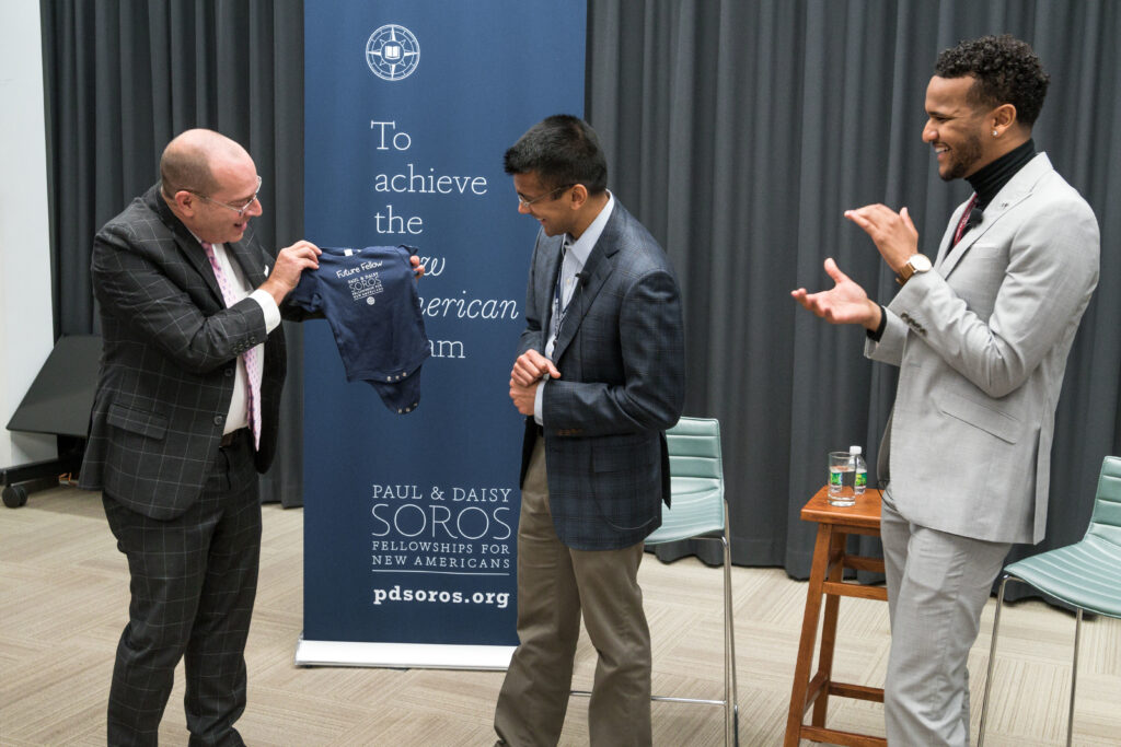 Photograph of a man dressing in a blue blazer and khaki pants. He is being presented a baby onesie from camera left and smiling. 