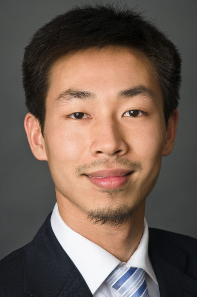 Headshot of a man in his 20s who has heritage from China, with medium skin tone, short black hair and a short mustache/goatee. He is wearing a black suit, white button up shirt and a blue striped tie. 