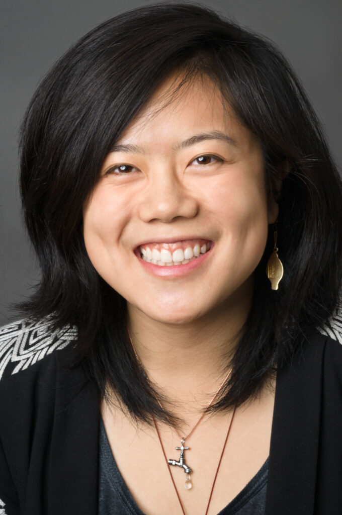 Headshot of a woman in her 20s who has heritage from China with light skin tone and shoulder length black hair with swoopy bangs. She is wearing a black jacket with white patterned accent shoulders, a grey shirt, a couple necklaces and dangly brass earrings. She is smiling at the camera.