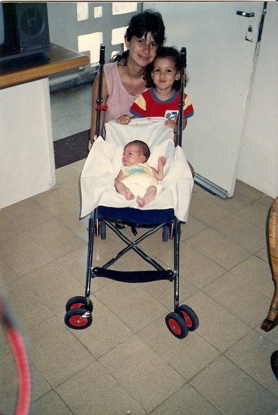 Old photograph of a woman in her 20s with light skin tone and loosely pulled back hair, she is crouched behind a small stroller with a baby, a young girl stands next to the woman. 