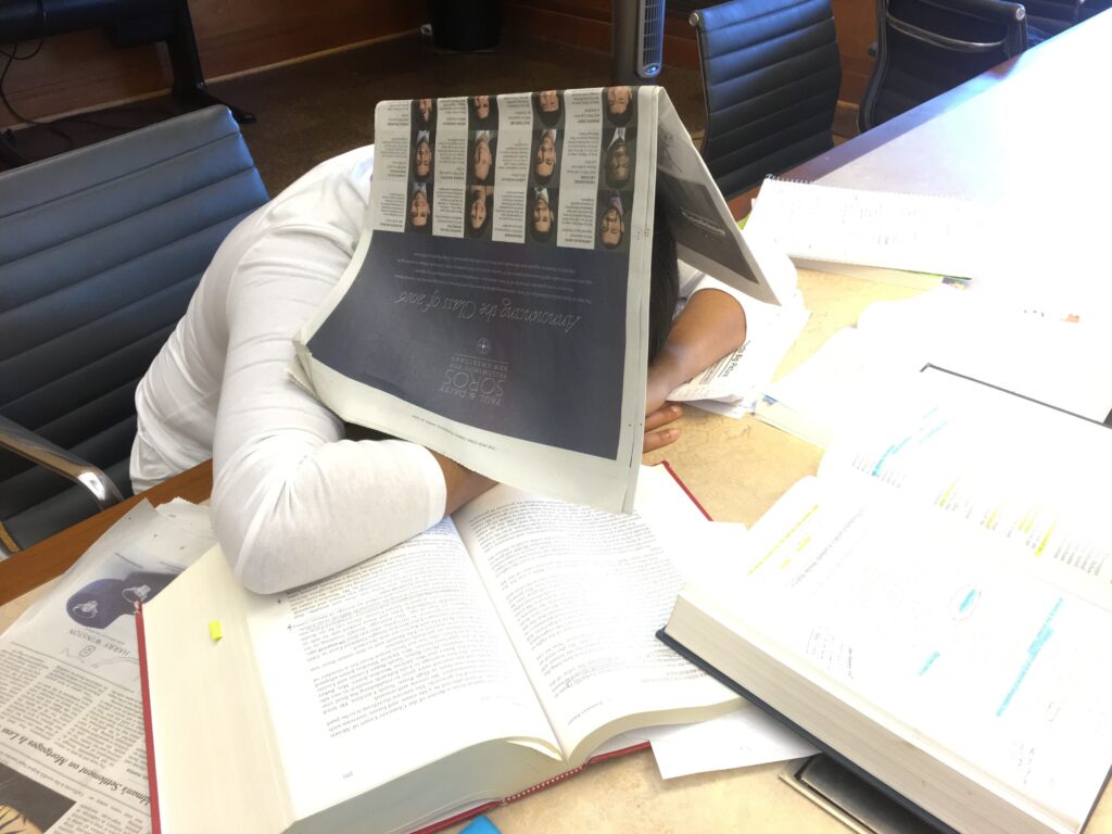 Photograph of a person resting their arms on a desk and their head on top of their hands, a New York Times is open resting on their head.