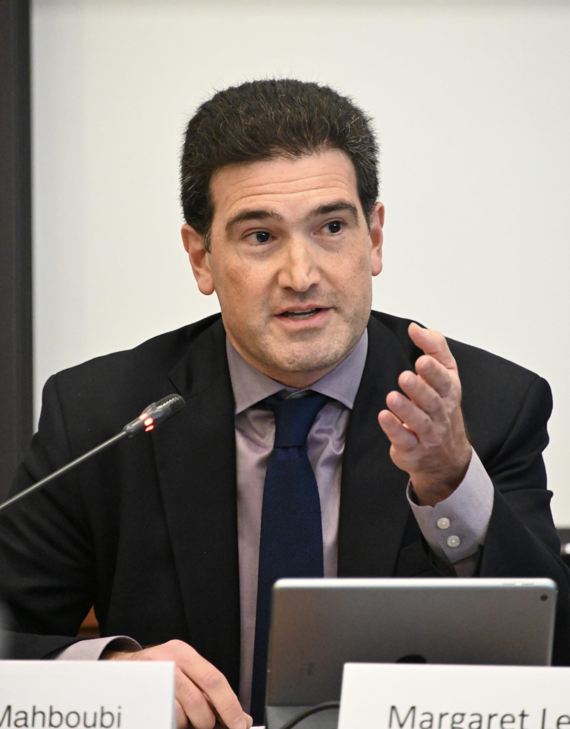 Photograph of a man sitting at a table with a microphone, iPad and name tag in front of him. He has heritage from Iran with light skin tone and black short hair. He is wearing a black suit, purple button up shirt and navy tie. He is looking off camera and appears to be speaking.