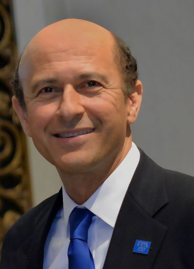 Headshot of a man in his 50s with light skin tone and brown balding hair. He is wearing a black suit and white button up. He is looking at the camera and smiling. 