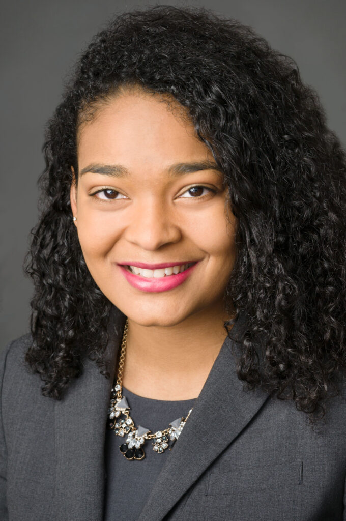 Headshot of a woman in her 20s with heritage from the Dominican Republic and has medium skin tone and curly black shoulder length hair with a right-side part. She is wearing a grey blazer with grey shirt and a black, grey and white jeweled necklace. She is smiling at the camera. 