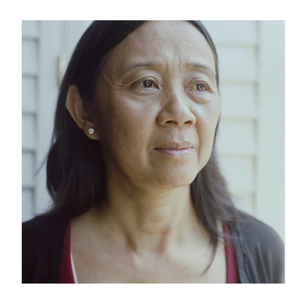 Photograph of a woman in her 40s who has light-medium skin tone and shoulder length black hair. She is wearing a black top over a red top, and stud earrings. There is a white slatted wall behind her. 