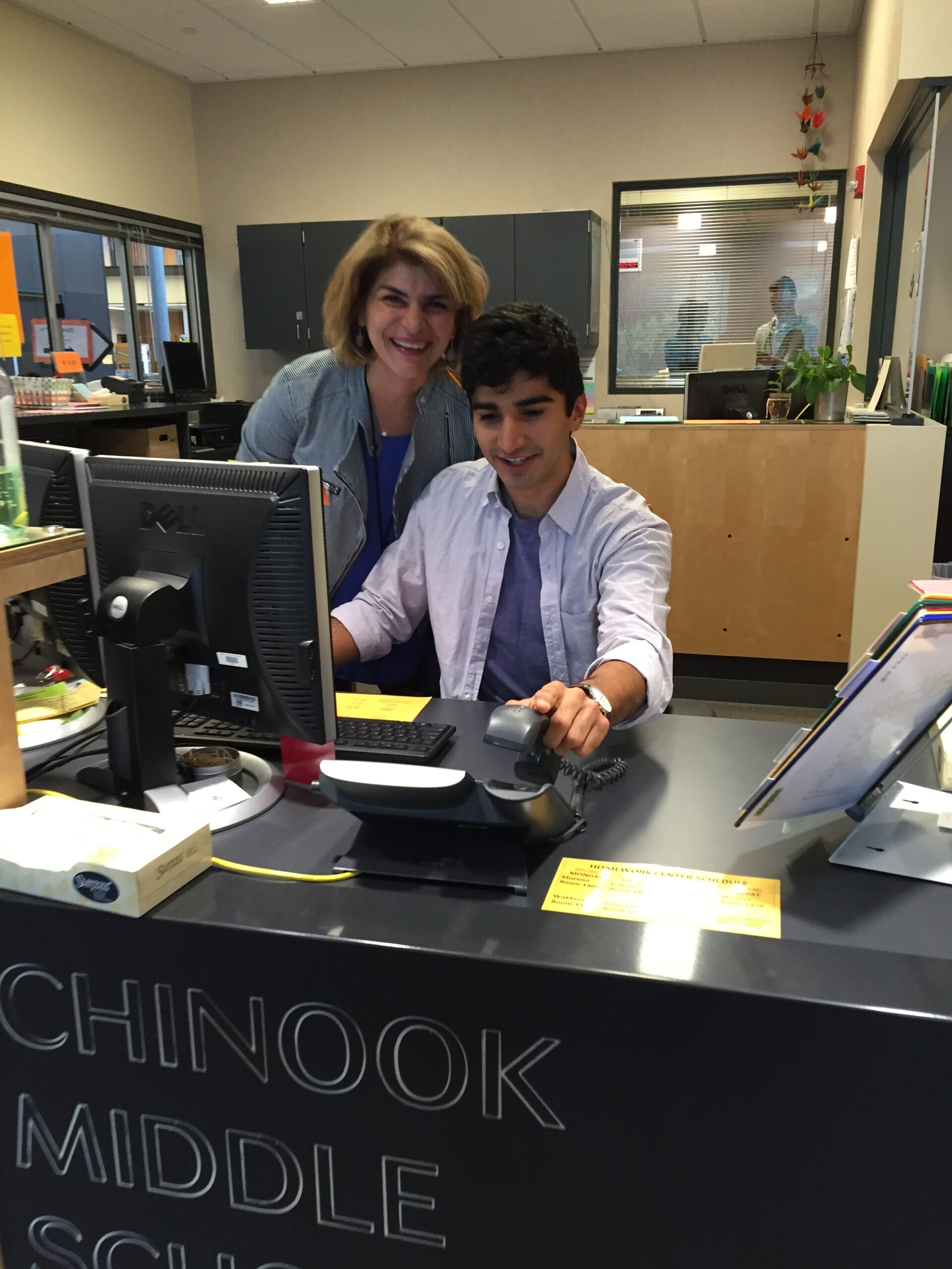 Photograph of a man in his 20s who has heritage from Iran with light skin tone and black hair, sitting behind a computer at a desk, a woman stands behind him looking on. They are both smiling. 