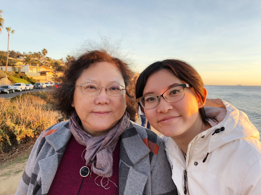 Photograph of two women, the road and large body of water can be seen behind them. The woman on the right is in her 20s with light skin tone and dark hair pulled back, she is wearing a white rain jacket and glasses. The woman on the left is middle-aged wearing a grey jacket, red top and glasses. 