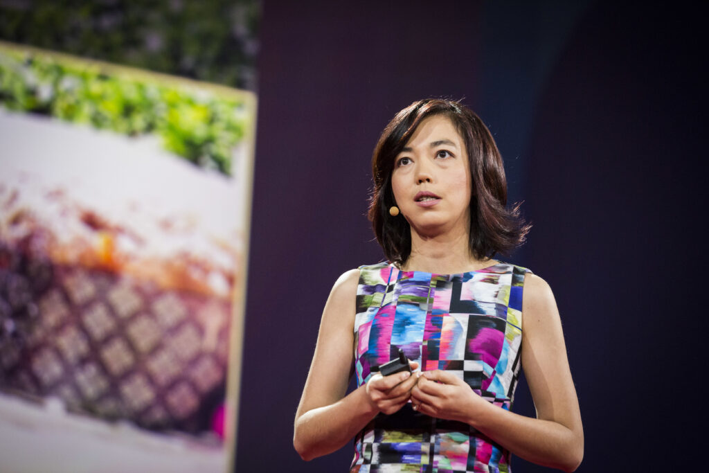 Photograph of a woman in her 40s who has heritage from China with light skin tone and shoulder length black hair. She is wearing a multi-colored sleeveless dress and a microphone can be seen next to her chin. She is holding a power point controller. 