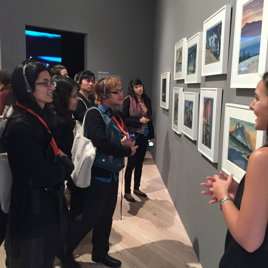 Photograph of a group of people having a variety of heritages, all in their 20s stands listening to headsets while looking at framed art. 