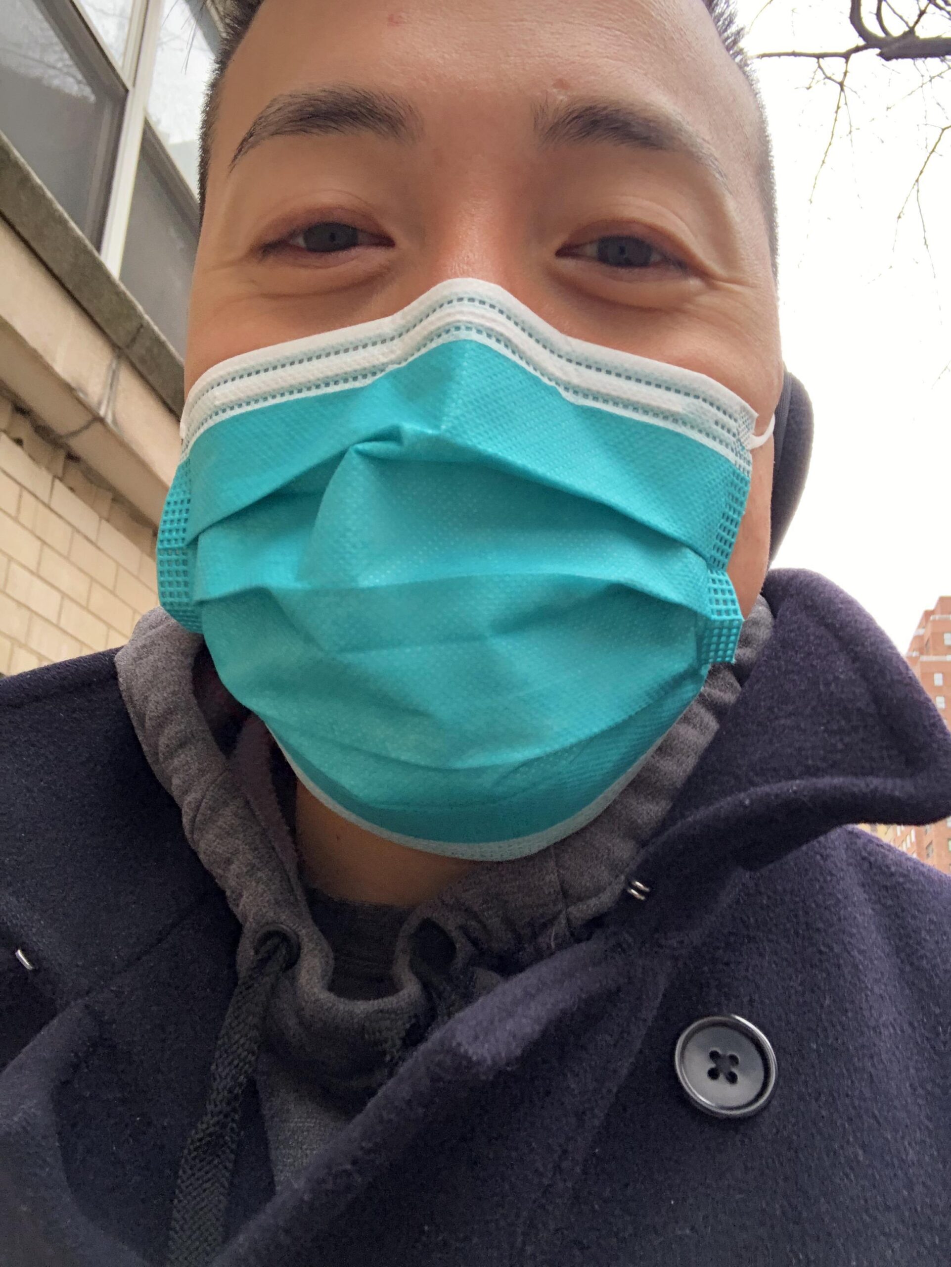 Selfie of a man in his 40s who has heritage from South Korea with light-medium skin tone and black hair. He is wearing a surgical mask, blue coat and grey sweatshirt.