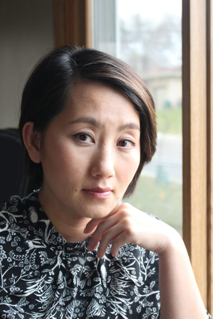 Headshot of a woman in her 30s who has heritage from Thailand, she has black hair with some highlights, cut short in the back and longer in the front. She is looking at the camera with her hand resting under her chin. She is wearing a black and white floral patterned top.