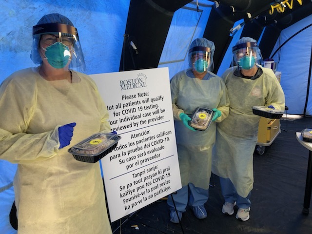 Photograph of three people standing, they are wearing all forms of PPE (protective personal equipment). They are holding to go containers of food. 