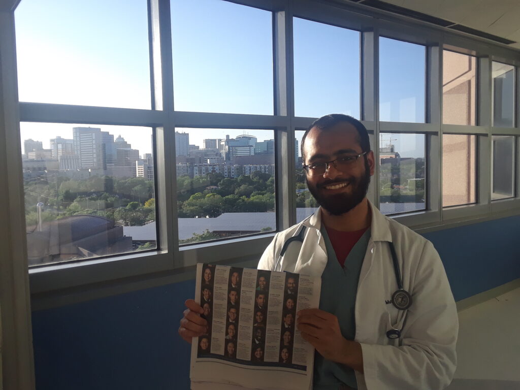 Photograph of a man in his 20s who has heritage from Pakistan with medium skin tone, buzzed black hair and short beard. He is wearing a white button up shirt and holding a New York Times. There are windows behind him, a body of water, trees and more buildings can be seen. 