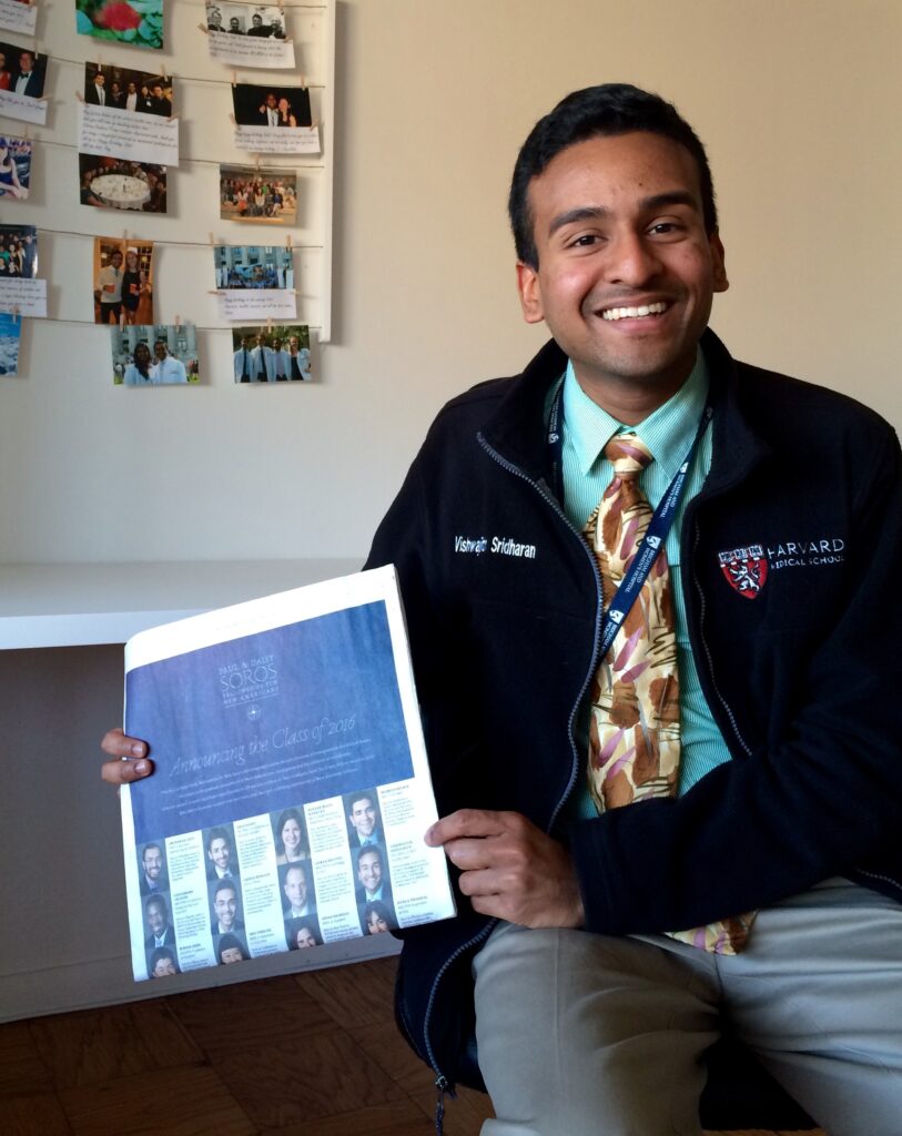 Photograph of a man in his 20s with medium skin tone and short black hair. He is wearing a fleece jacket, teal button up and multi-colored tie. He is holding a New York Times and smiling.