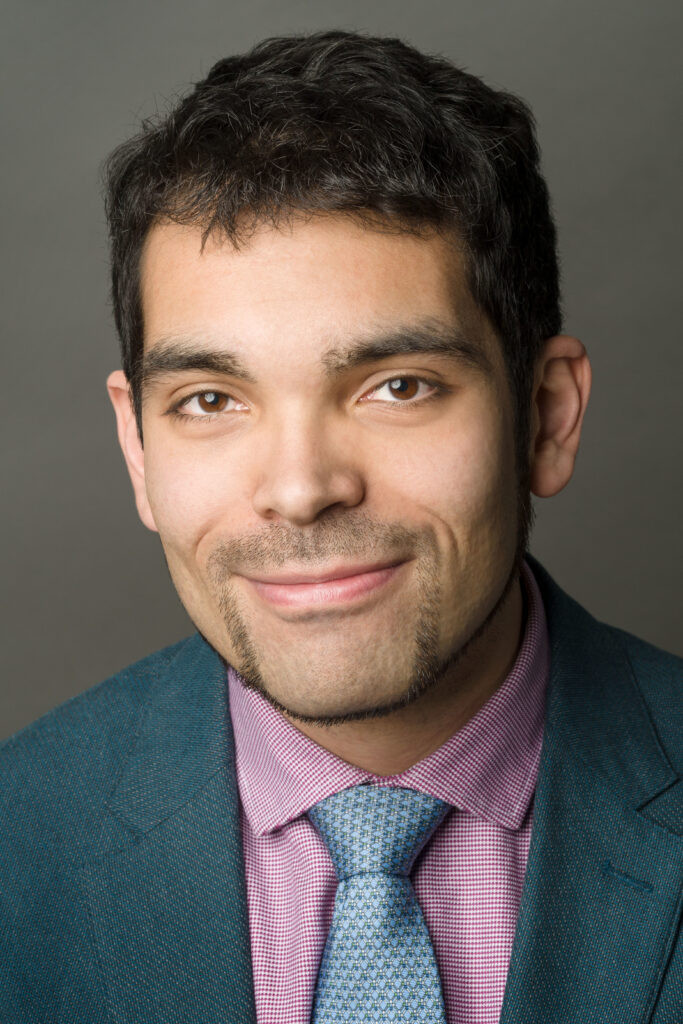 Headshot of a man in his 20s who has heritage from Mexico and Colombia with light skin tone, short black wavy hair and a goatee. He is wearing a dark teal blazer and red and white gingham button up and blue tie. He is smiling at the camera. 