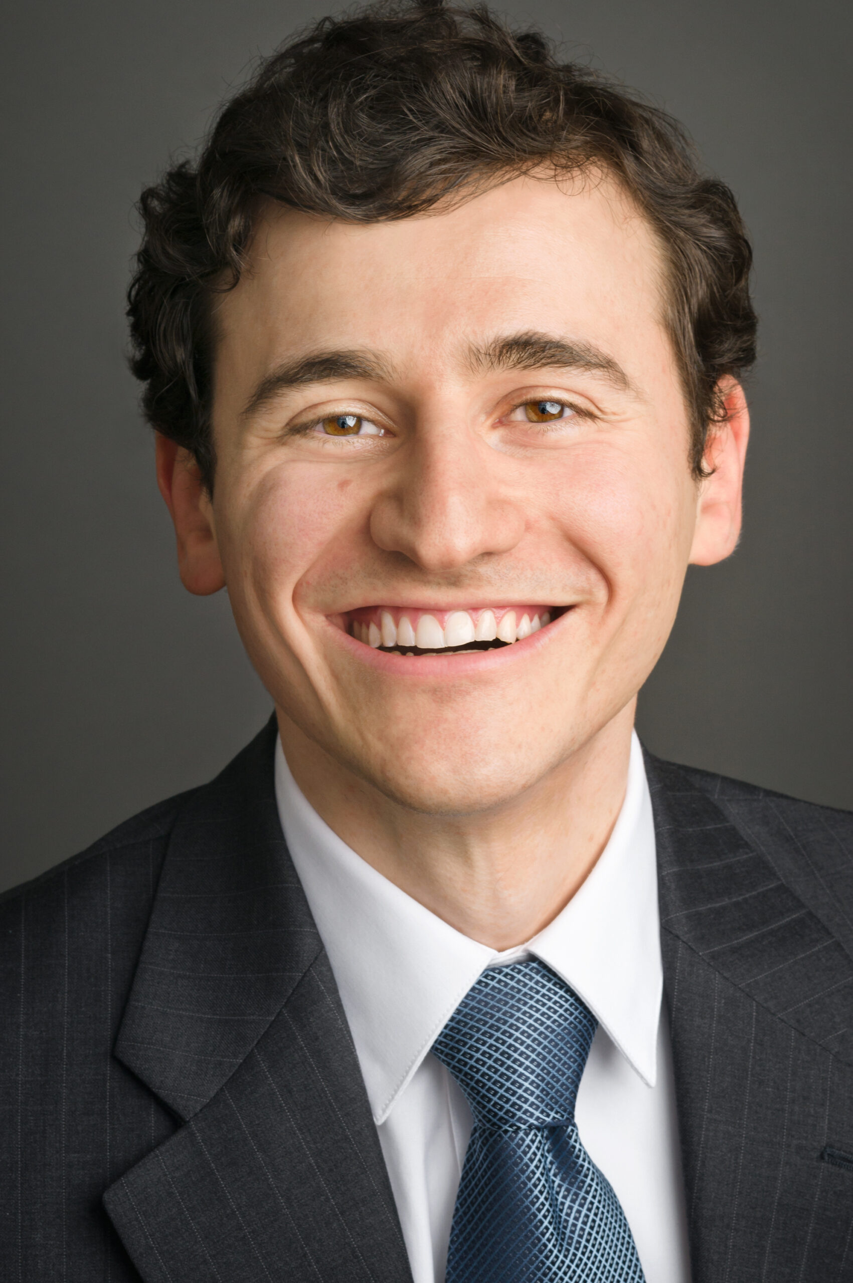 Headshot of a man in his 20s who has heritage from Ukraine with light skin tone and dark brown wavy hair cut short. He is wearing a grey pinstripe suit, white button up shirt and blue tie. He is smiling at the camera.