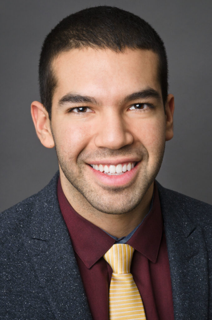 Headshot of a man in his 20s who has heritage from El Salvador with light skin tone and black buzzed hair. He is wearing a navy speckled suit, maroon button up and yellow tie. He is smiling at the camera.