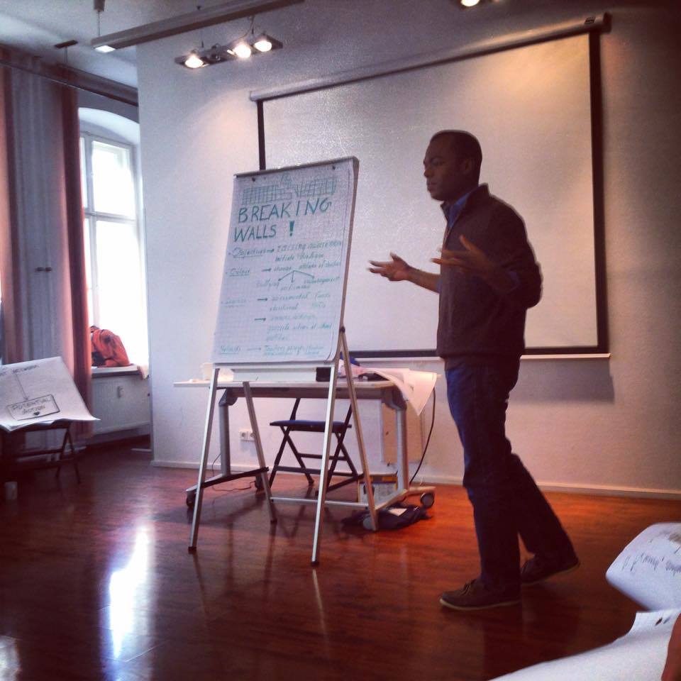 Photograph of a man standing at the front of a room and gesturing with his hands.