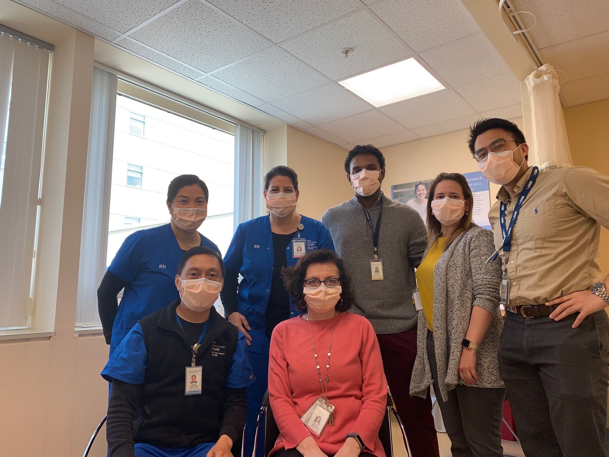 Photograph of a group of people standing and posing for the camera. They are in a hospital room and all wearing surgical masks. 