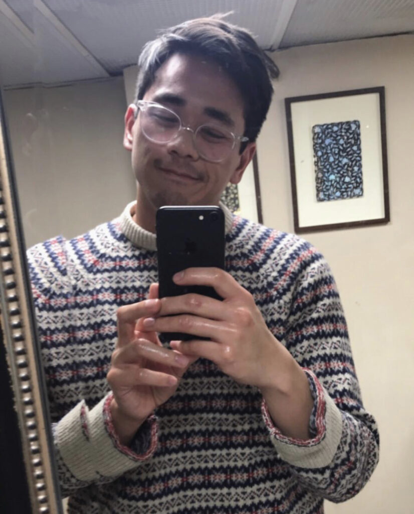 Mirror selfie of a man in his 20s wearing a white, blue and red snowflake style pattern sweater. 