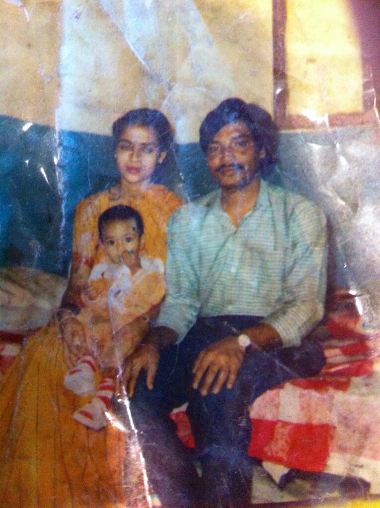 Old, wrinkled photograph of a family, mom is in her 20s and has light skin tone and pulled back black hair, she is wearing a marigold saari and holding a baby, also in marigold and white. Dad in his 20s and has medium skin tone and black hair, with a mustache, he is also sitting and wearing a green patterned button up shirt, navy slacks.