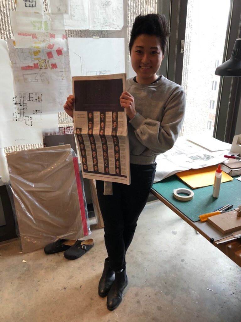 Photograph of a woman in her 20s who has heritage from South Korea with light skin tone and pixie short black hair. She is standing near a worktable; paper tape and glue can be seen. She is holding a New York Times. 