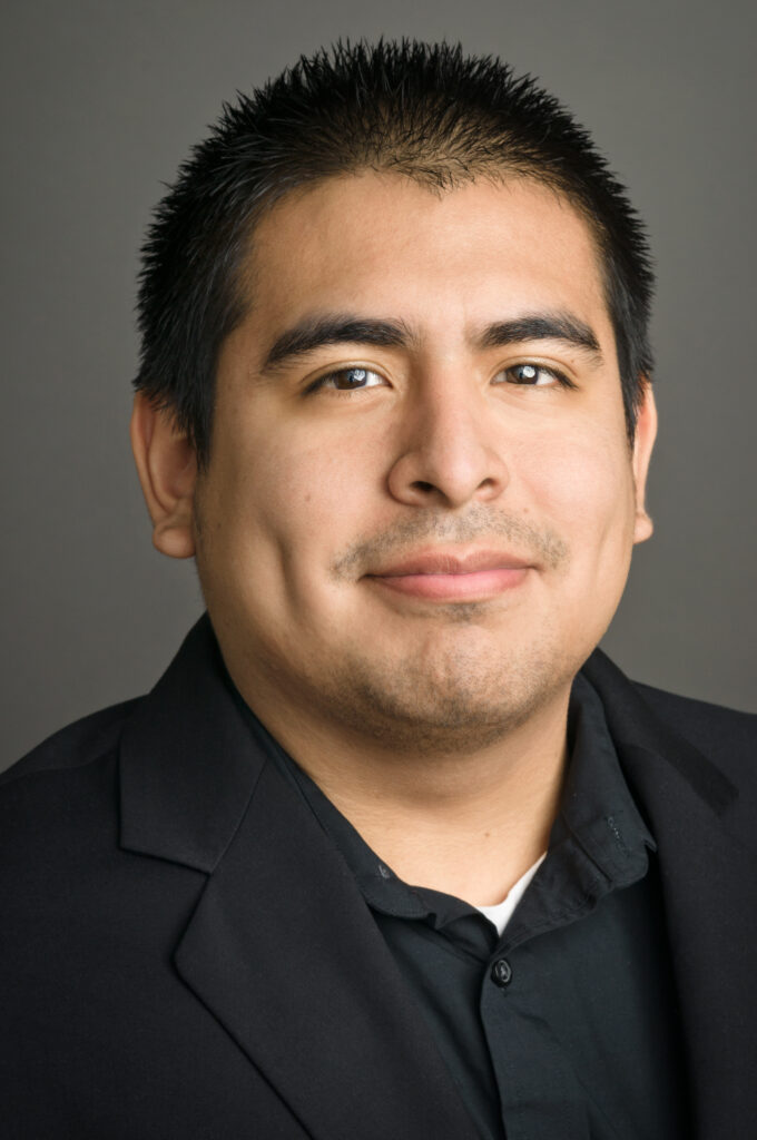 Headshot of a man in his 30s who has heritage from Peru with light skin tone and black short hair. He is wearing a black blazer and black button up and smiling at the camera.