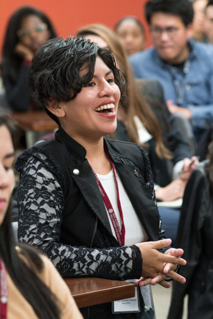 Photograph of a group of people, one woman is in focus, with light skin tone and short black hair, she is wearing a white shirt and black jacket with lace sleeves on top. She is smiling and looking off camera. 