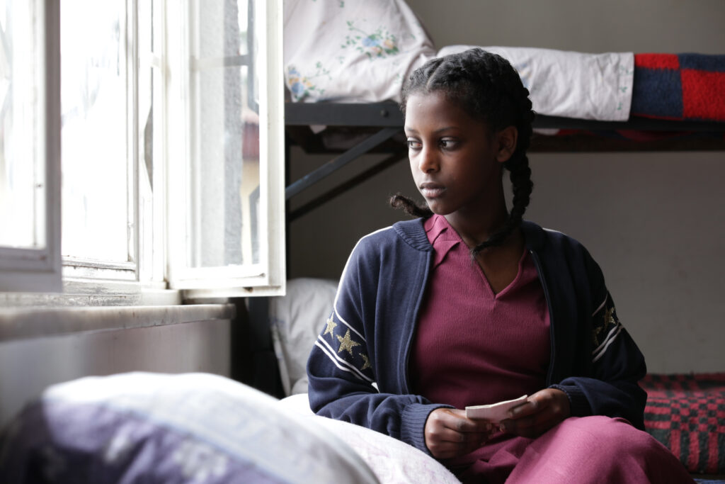 Photograph of a woman in her teens, she has dark skin tone and black hair in two French braids. She is wearing a red dress, and navy long sleeve with gold stars on the arms. She looks out a window to her right, there are bunk beds made with blankets behind her.