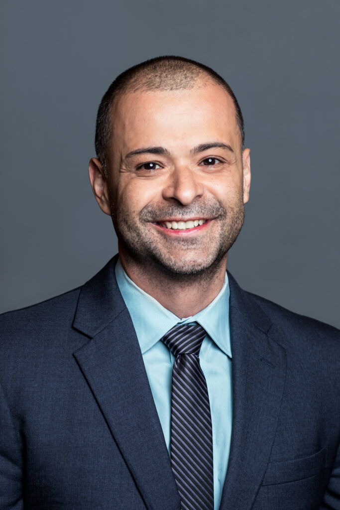 Headshot of a man in his 30s who has heritage from Colombia with light skin tone and buzzed black hair, with a 5-o'clock shadow beard. He is wearing a navy suit, light teal button up and navy striped tie. He is smiling at the camera. 