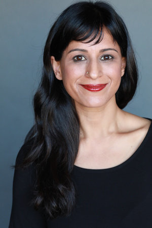 Headshot of a woman in her 30s who has heritage from India. She has light-medium skin tone and long, slightly wavy black hair and bangs. She is wearing a black scoop neck top, and has a stud in her right nostril. She is looking at the camera and smiling.