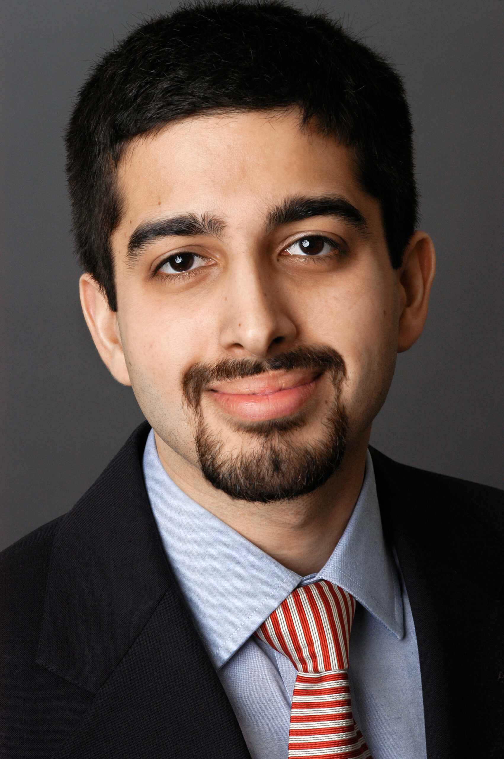 Headshot of a man in his 20s who has heritage from India with light skin tone, short black hair and a goatee/ mustache. He is wearing a black suit, light blue button up shirt and red and white striped tie. He is smiling at the camera. 