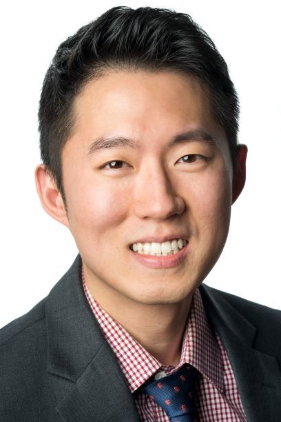 Headshot of a man in his 20s who has heritage from South Korea, he has light skin tone and black hair. He is wearing a dark grey suit, red and white gingham button up and navy tie. He is smiling at the camera.