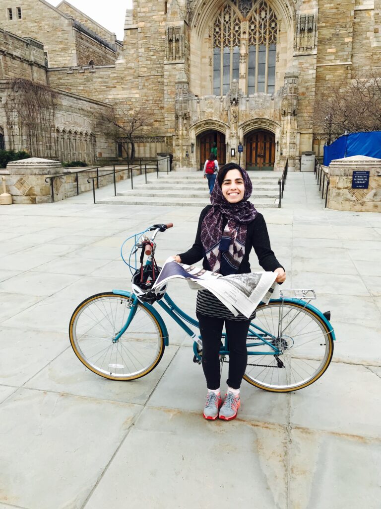 Photograph of a woman in her 20s who has heritage from Afghanistan with light skin tone and black hair is seen peeking out from her dark blue hijab. She is standing next to a teal bicycle holding a New York Times. A large ornate brick building can be seen behind her.