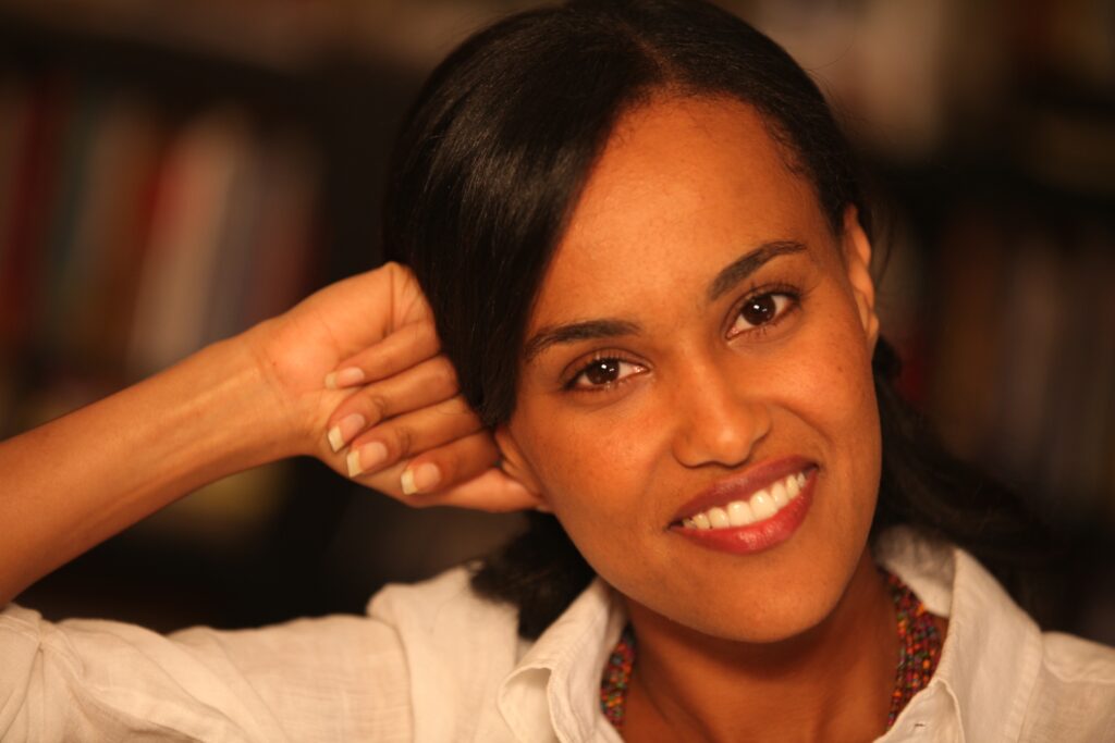 Headshot of a woman in her 20s who has heritage from Ethiopia with medium skin tone and black pulled back hair. She is wearing a white button up shirt. She is leaning against one fist and smiling at the camera. 