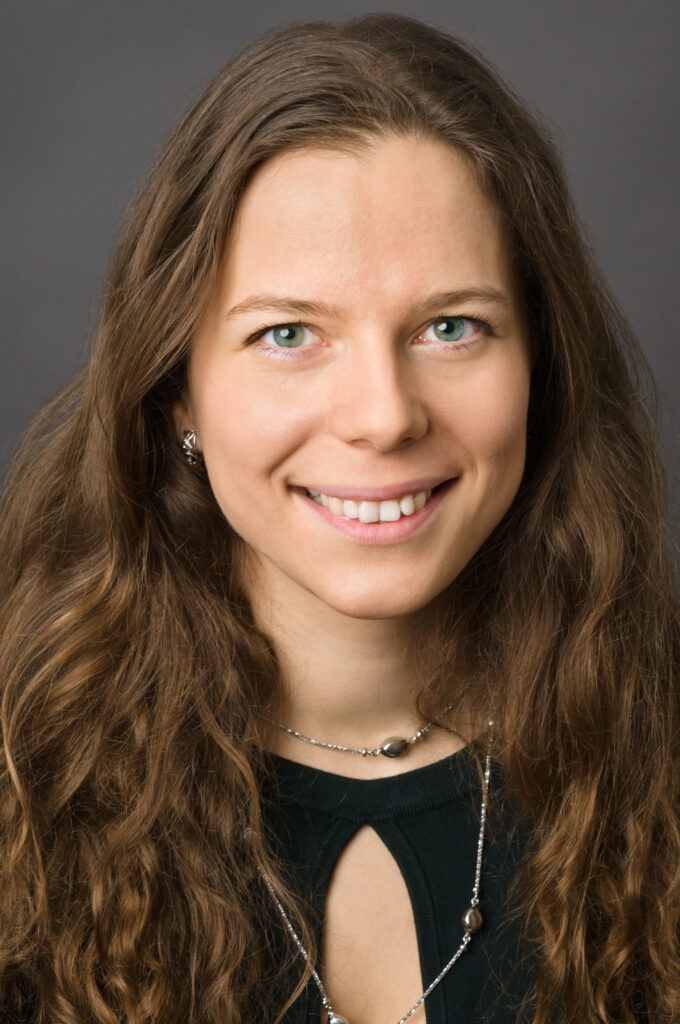 Headshot of a woman in her 20s who has heritage from Russia with light skin tone and long wavy light brown hair parted in the middle. She is wearing a black top with a keyhole, long silver necklace and silver earrings. She is smiling at the camera.