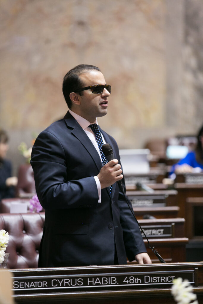 Photograph of a man in his 30s who has heritage from Iran with light skin tone and short black hair, he is wearing a grey suit, light pink shirt and blue tie with dark sunglasses. He is holding a microphone and there is a sign at his desk that says "Senator Cyrus Habib, 48th District"