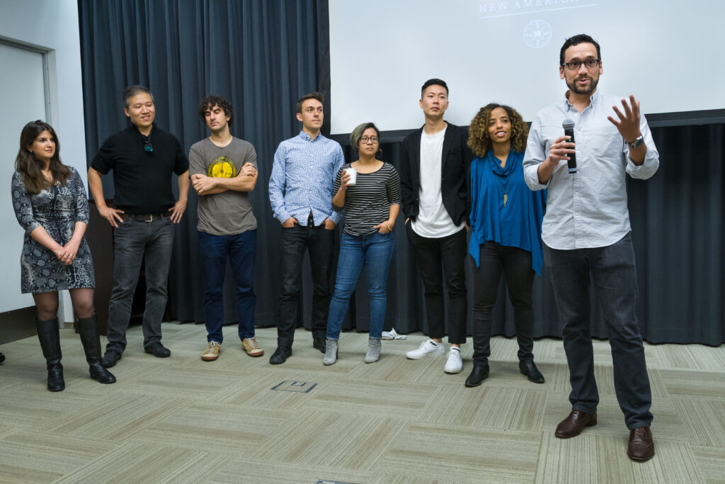 Photograph of eight people standing with a screen behind them, they are of varying genders, ages and heritages. One person has a microphone in hand and is speaking.