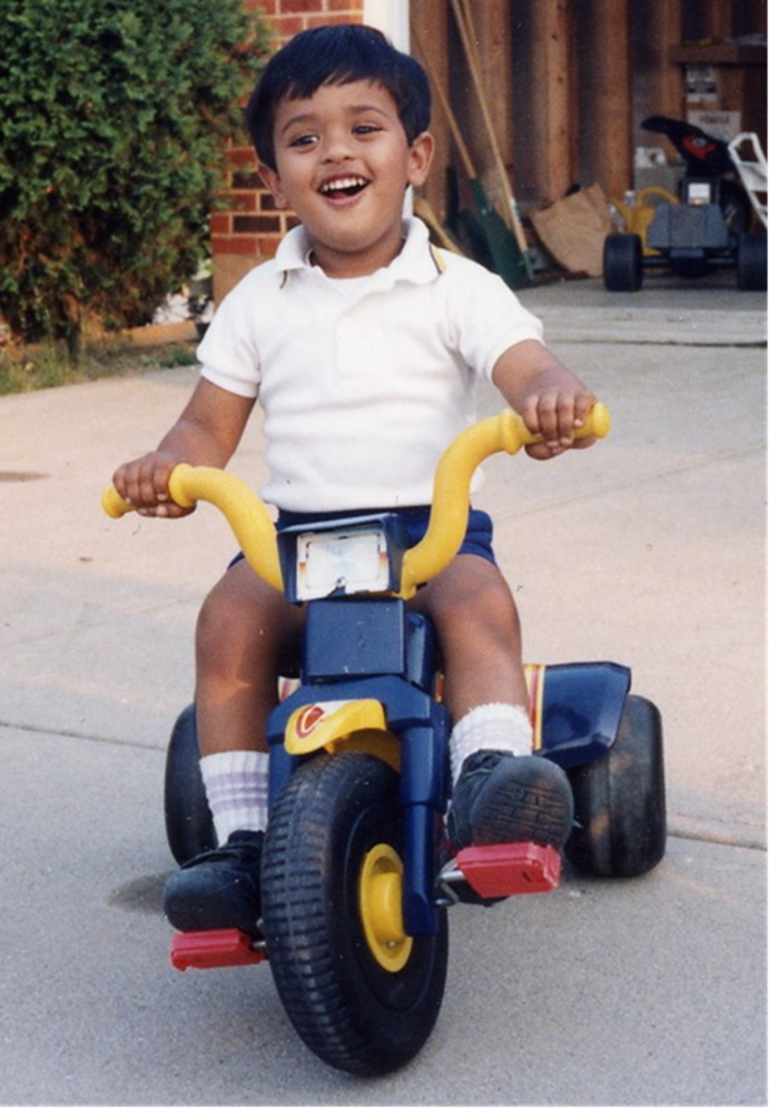 Old photograph of a young boy with medium skin tone and black hair, wearing a white polo and blue shorts rides ad tricycle and laughs. 