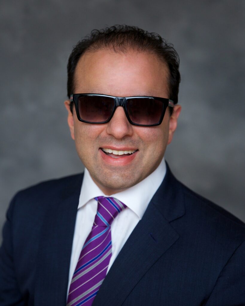 Headshot of a man in his 30s who has heritage from Iran with light skin tone and black short hair, combed back. He is wearing a navy suit, white button up and purple and blue striped tie, and large square sunglasses. He is smiling at the camera. 